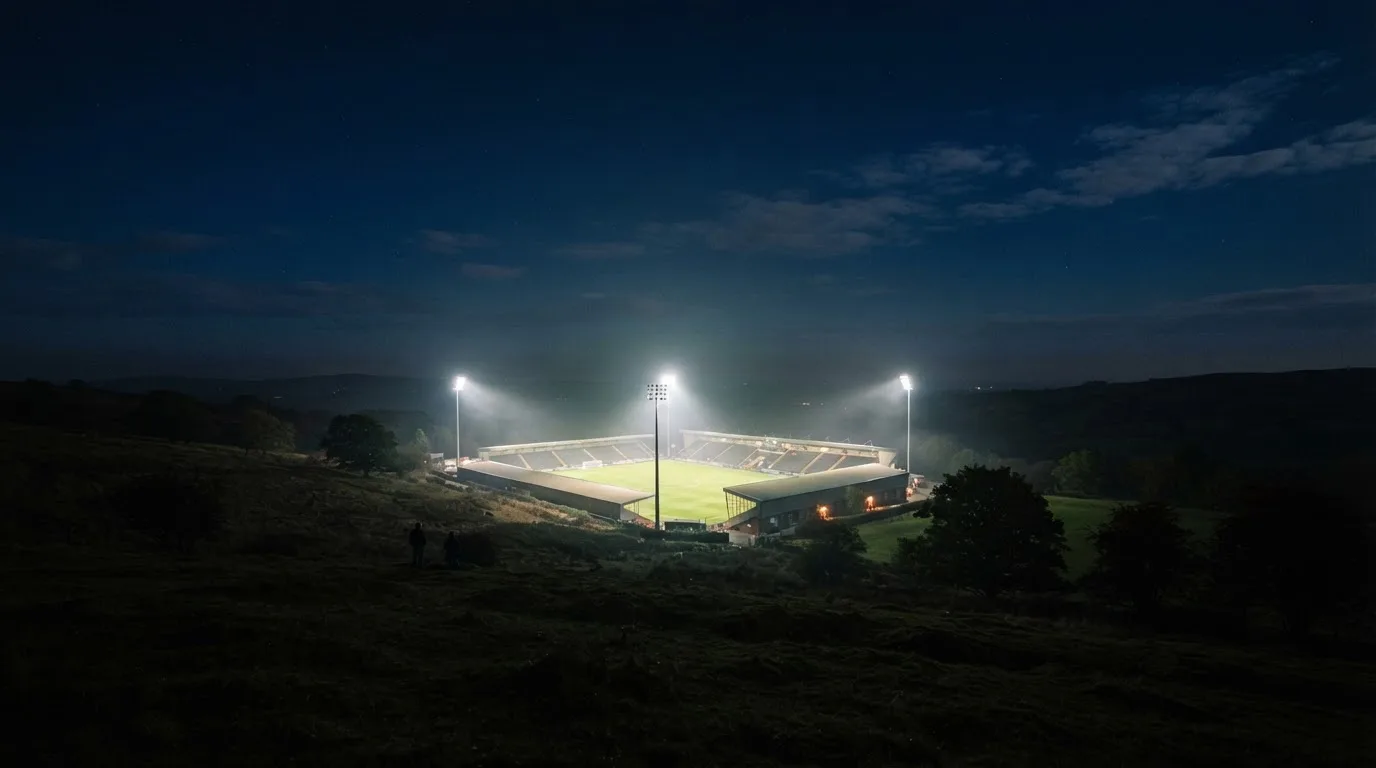 Ein Stadion unter Flutlicht bei Nacht, von einem Hügel aus der Ferne betrachtet, mit Dunst über dem Spielfeld.