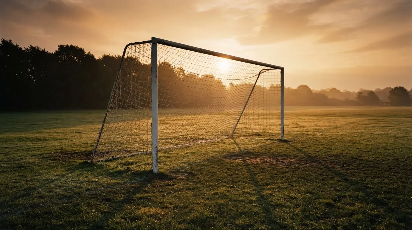 Ein Fußballtor mit Netz bei Sonnenuntergang auf einem leeren Platz, die Schatten der Torpfosten liegen lang auf dem Gras.