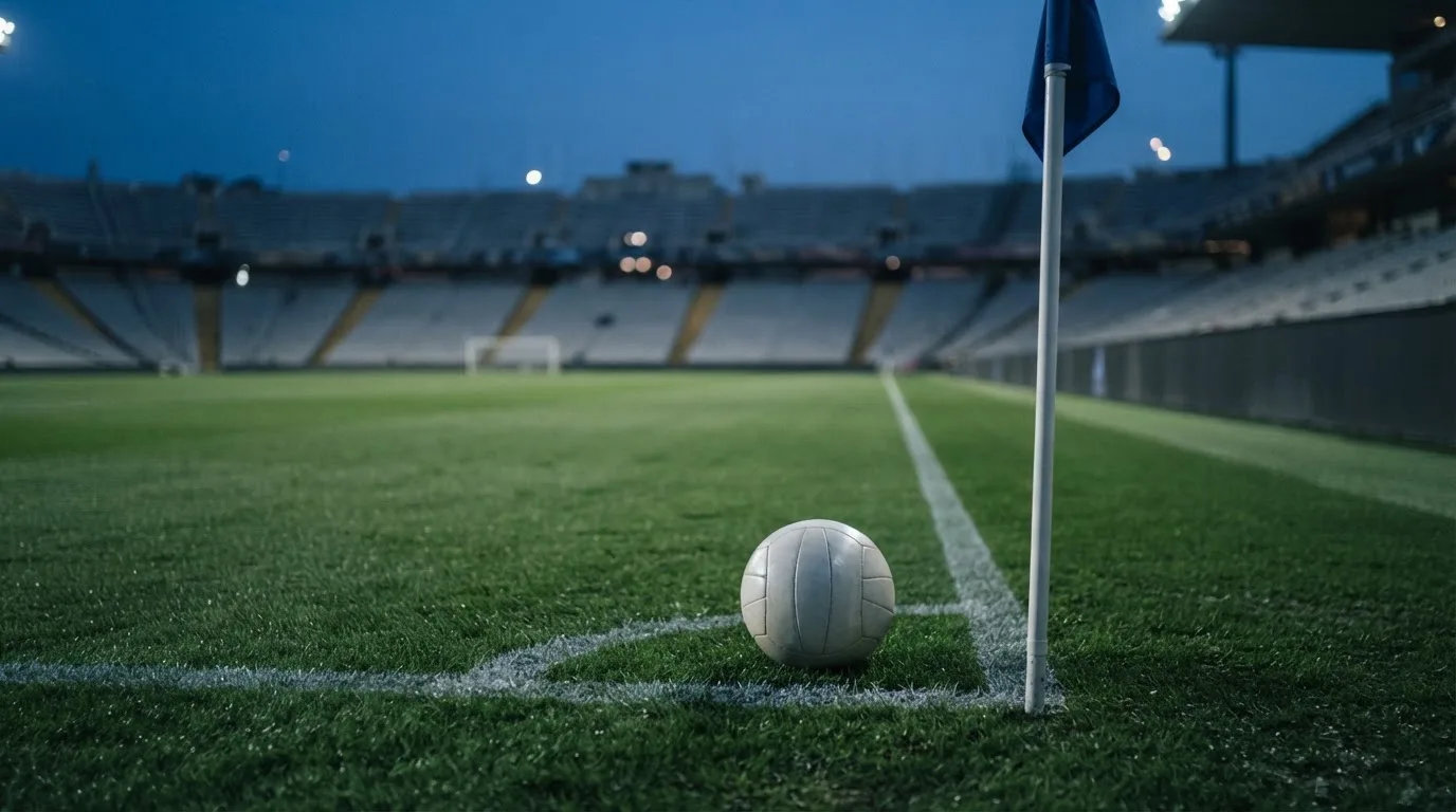 Ein Fußball liegt neben einem Eckfahnen-Pfosten auf einem Rasenplatz, im Hintergrund ein leeres Stadion in der Dämmerung.