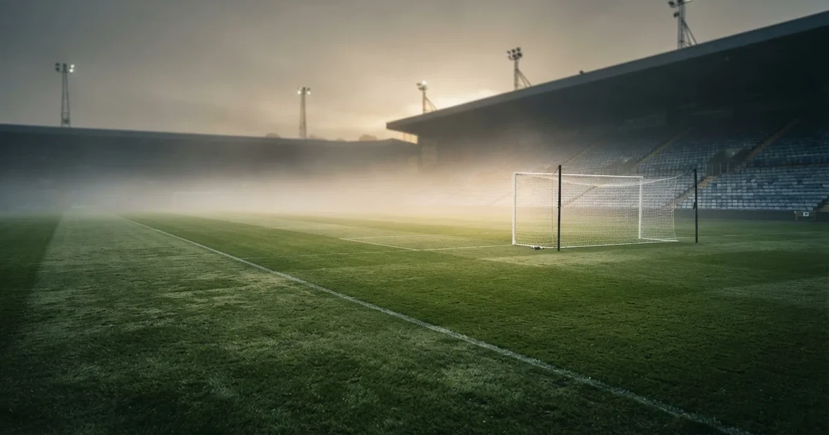 Ein Fußballplatz mit gepflegtem Rasen und einem Tor, über dem Spielfeld liegt Morgennebel, im Hintergrund eine leere Tribüne.