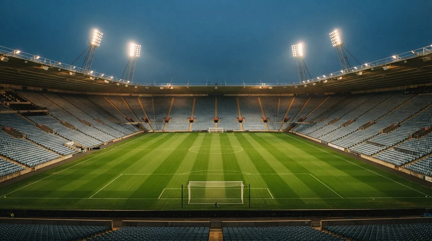 Eine leere Stadiontribüne unter Flutlicht aus der Ferne betrachtet, die Rasenfläche leuchtet grün in der Dämmerung.