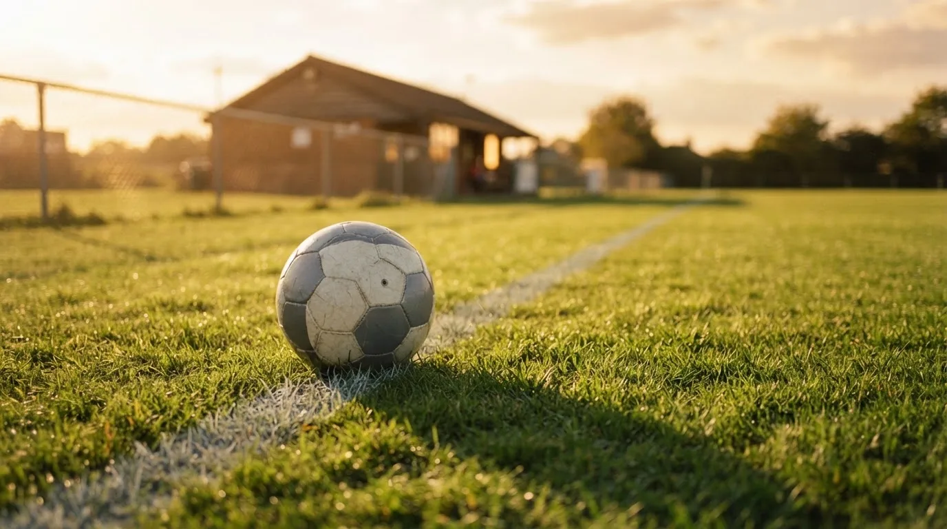 Ein Fußball liegt am Rand eines Rasens neben einer weißen Linie, im Hintergrund ein Zaun in der Abendsonne.