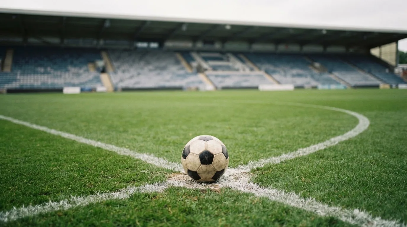 Ein Fußball liegt auf dem Anstoßpunkt eines leeren Rasenplatzes, im Hintergrund eine ruhige Tribüne unter bedecktem Himmel.