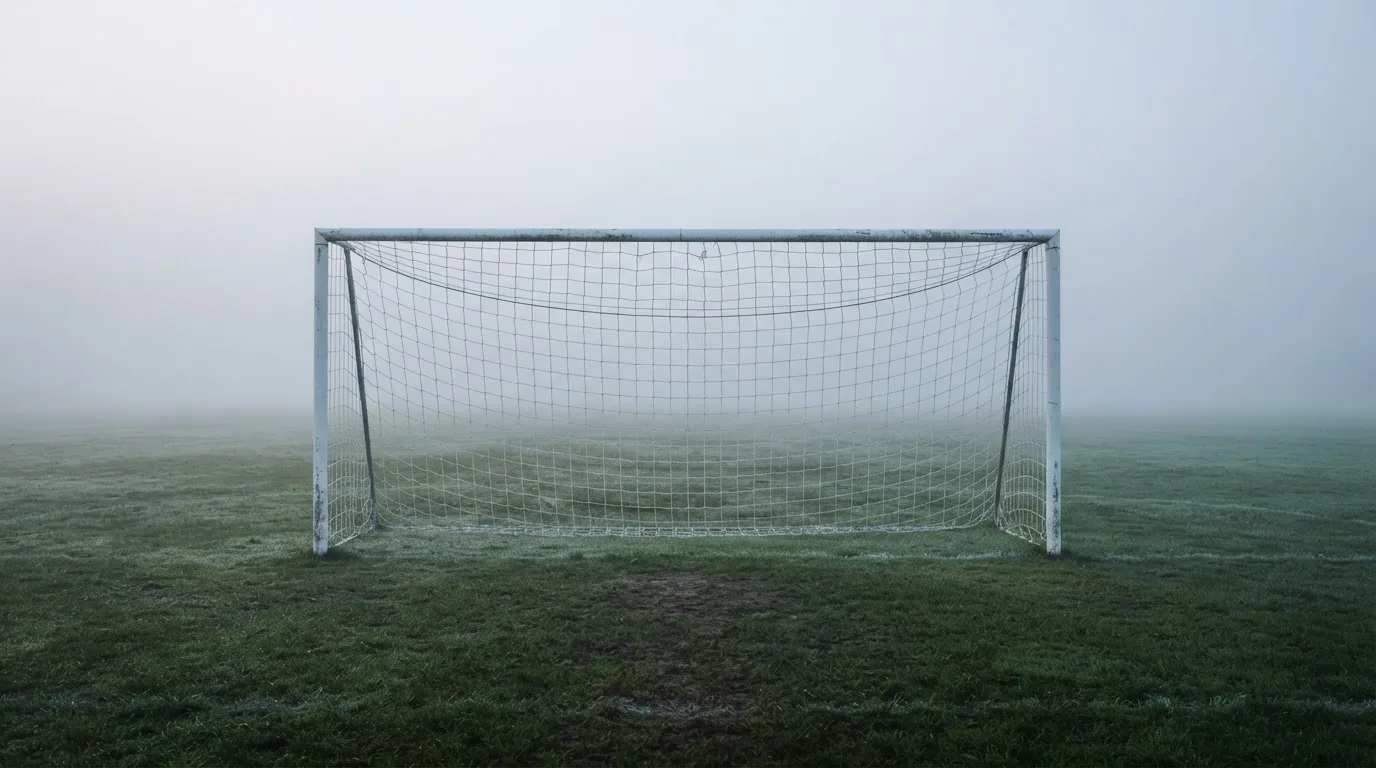 Ein Fußballtor mit Netz im Morgennebel auf einem leeren Platz, die Linien nur schwach zu erkennen.