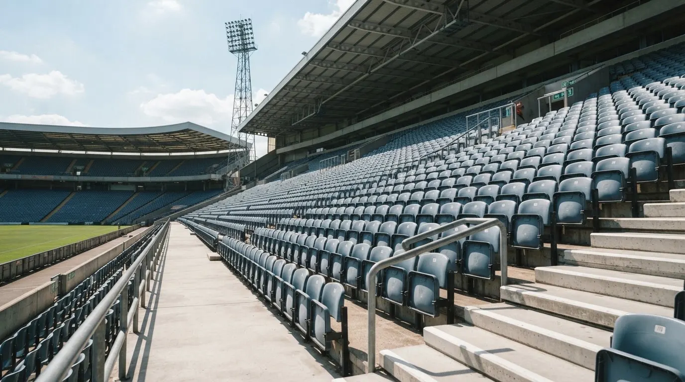 Eine leere Tribüne eines Fußballstadions bei Tageslicht, Sitzschalen in Reihen und dahinter ein Flutlichtmast.