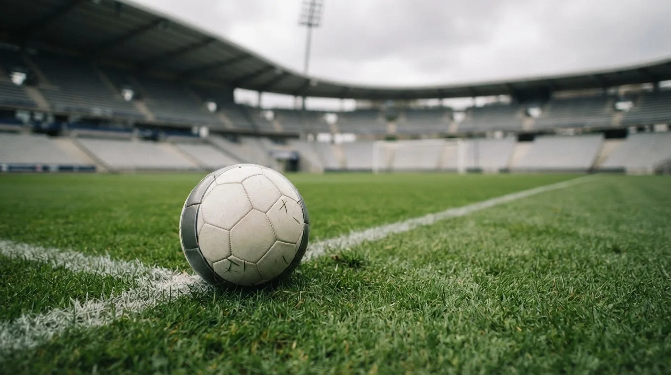 Ein Fußball auf einem Rasen, im Hintergrund ein leeres Stadion bei bewölktem Himmel.