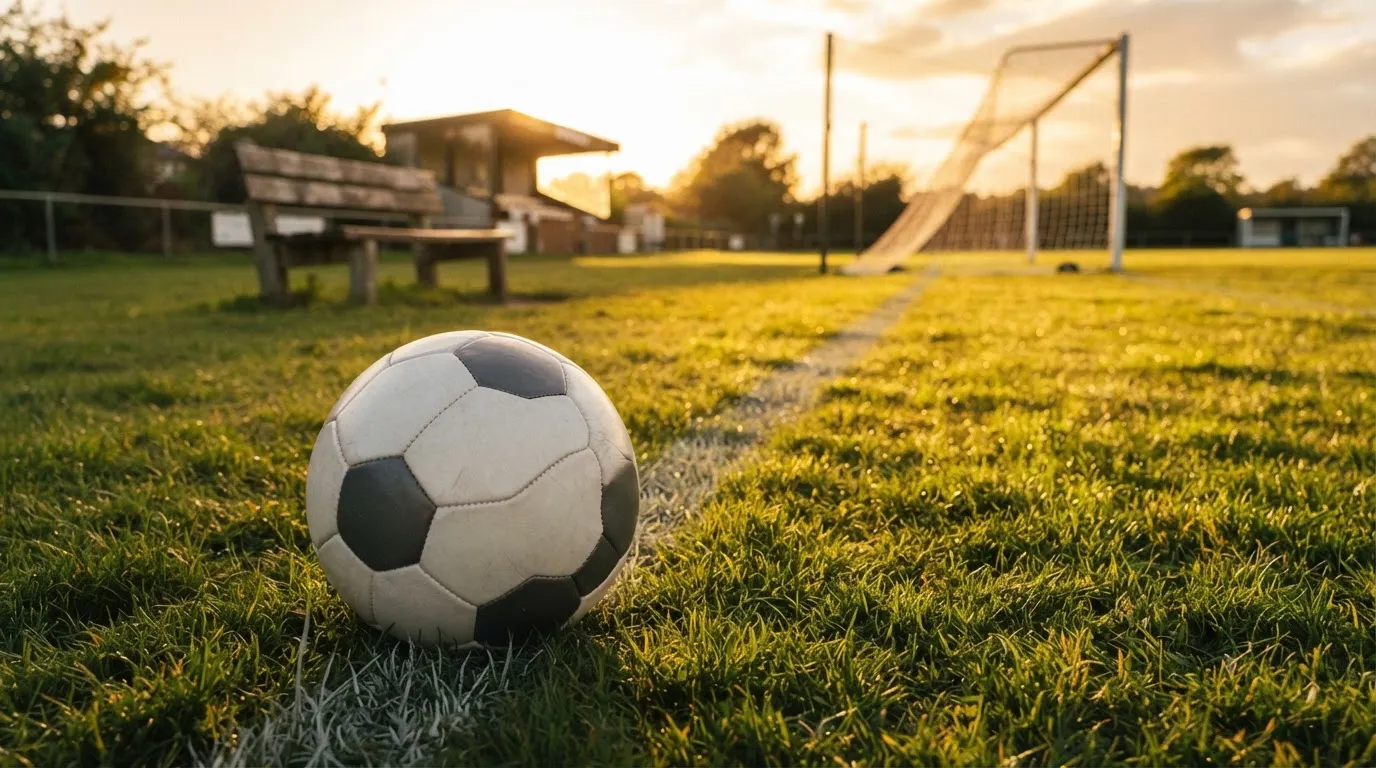Ein Fußball liegt im Gras am Spielfeldrand, dahinter eine leere Bank und ein Tor im Gegenlicht.