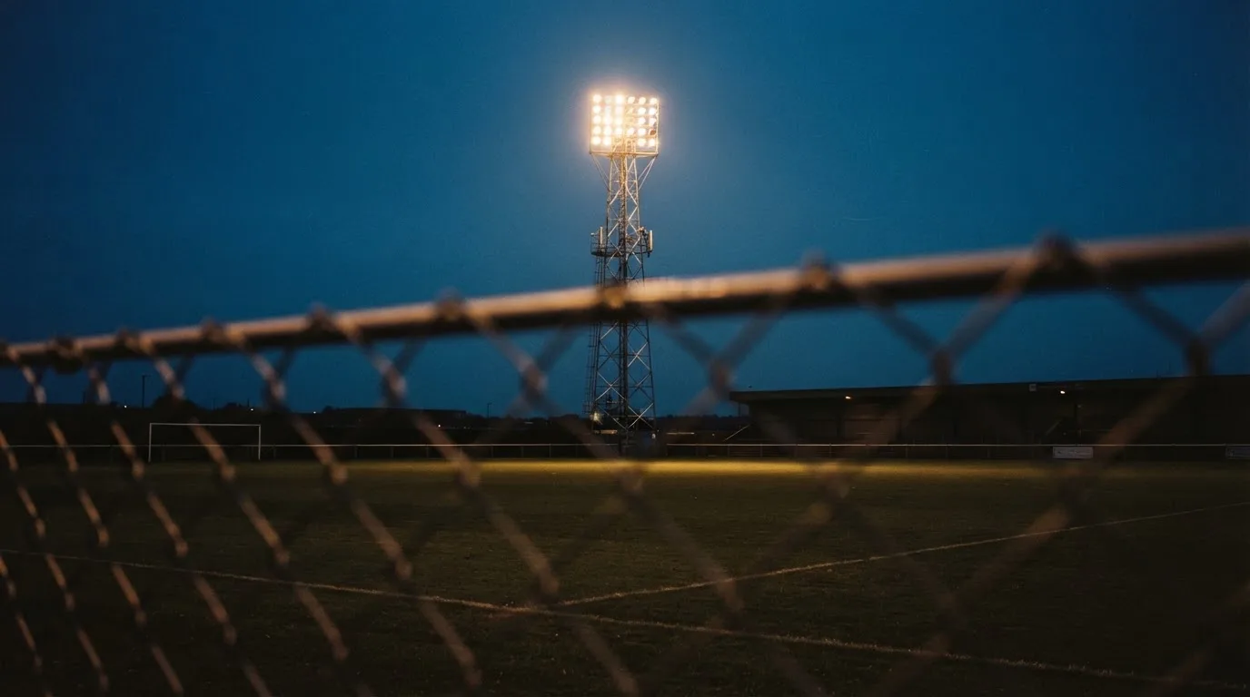 Eine Flutlichtanlage über einem leeren Sportplatz am Abend, im Vordergrund ein Maschendrahtzaun.