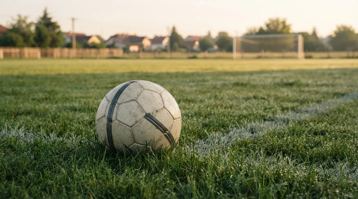Ein Fußball auf dem Rasen eines leeren Sportplatzes am Rand einer Kleinstadt im Morgenlicht.