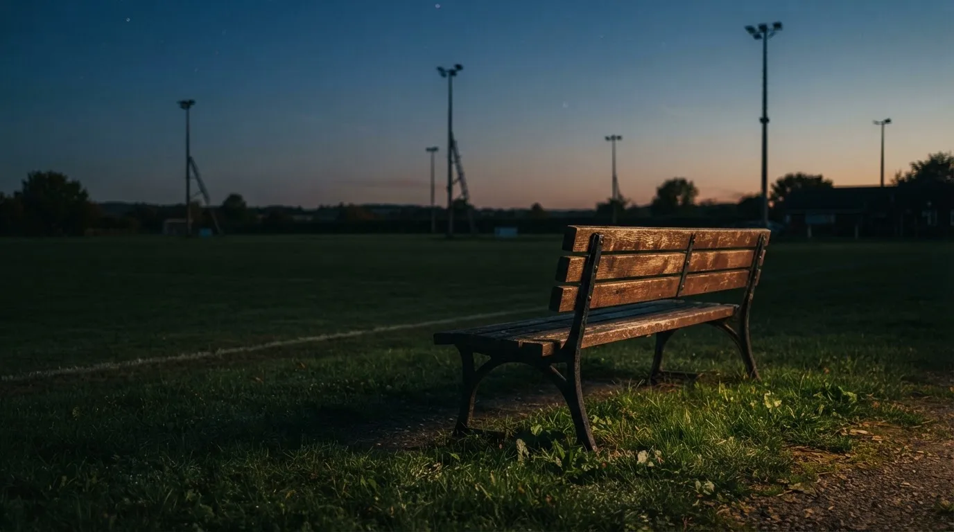 Eine leere Parkbank am Rand eines Fußballfelds in der Abenddämmerung, im Hintergrund ragen dunkle Flutlichtmasten in den Himmel.