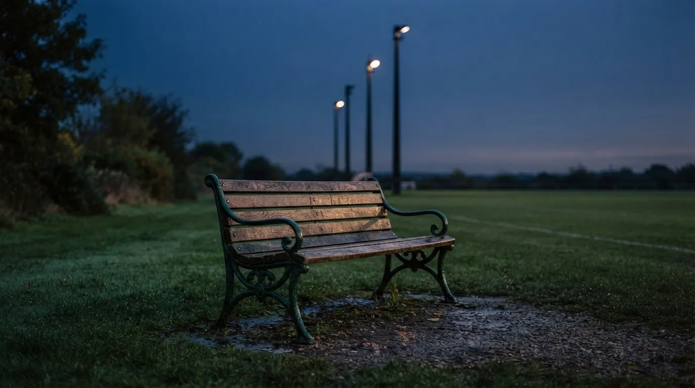 Eine leere Parkbank am Rand eines Fußballfelds in der Abenddämmerung, im Hintergrund ragen dunkle Flutlichtmasten in den Himmel.