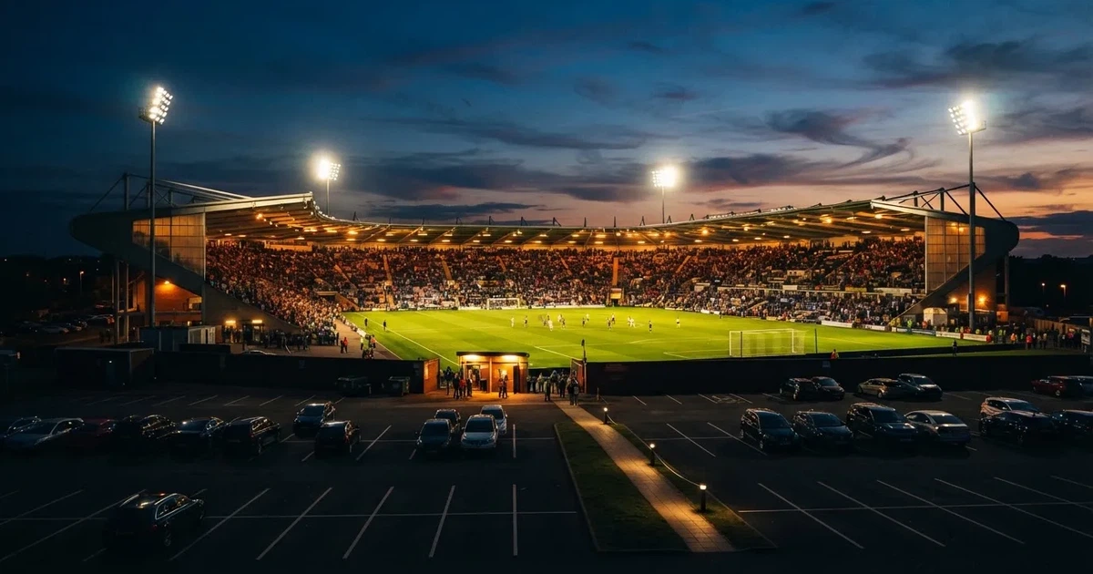 Ein Fußballstadion unter Flutlicht aus großer Entfernung, davor ein dunkler Parkplatz und ein heller Abendhimmel.