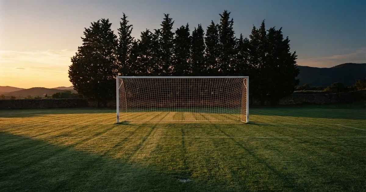 Ein Fußballtor mit Netz auf einem leeren Platz im Abendlicht, der Rasen frisch gemäht, im Hintergrund Zypressen.