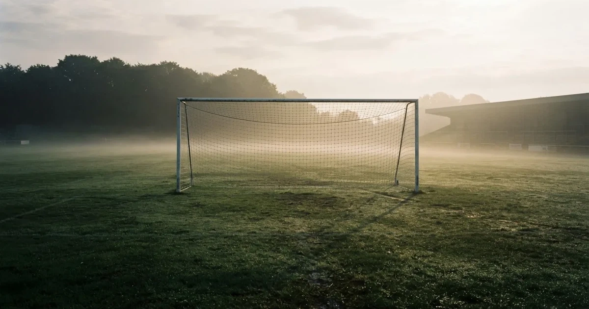 Ein Fußballtor mit Netz steht auf einem leeren Platz im Nebel der Morgendämmerung, der Rasen ist taufeucht.