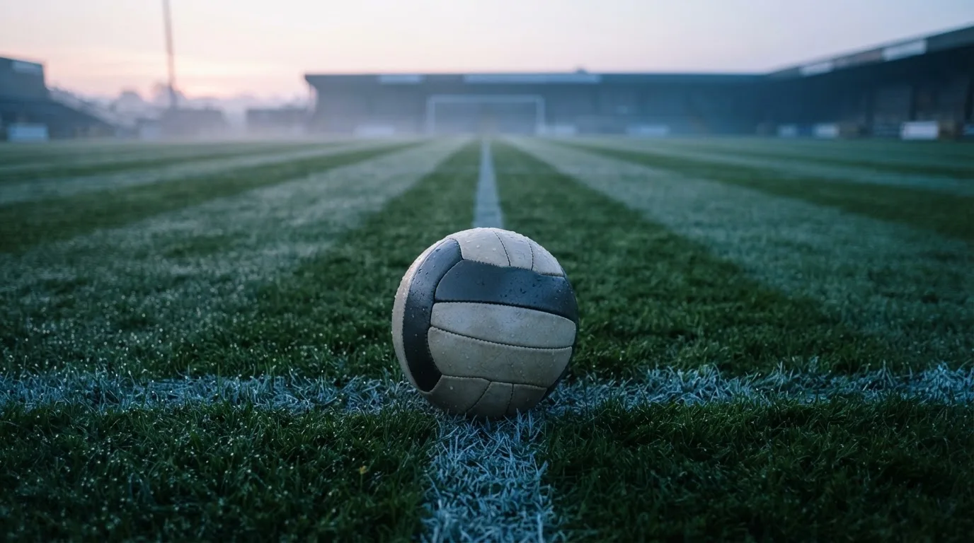 Ein Fußball ruht auf dem Mittelkreis eines leeren Rasenplatzes in der Morgendämmerung, Tau liegt auf dem Gras.