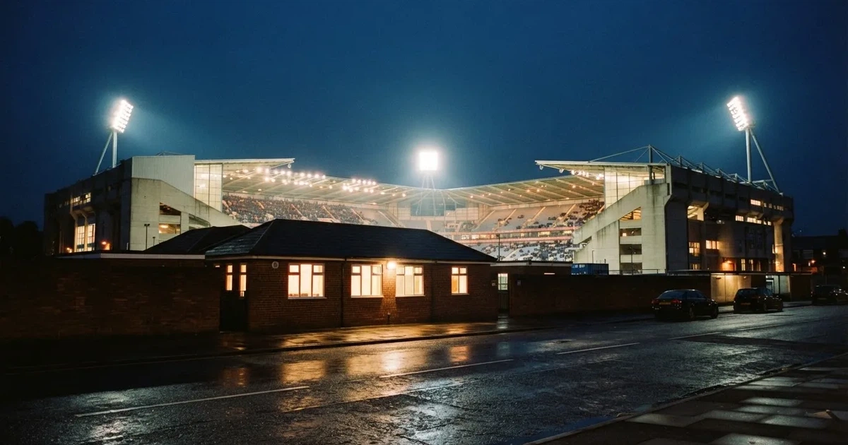 Ein Fußballstadion bei Flutlicht aus der Ferne gesehen, davor eine Straße und leuchtende Fenster eines Nebengebäudes.