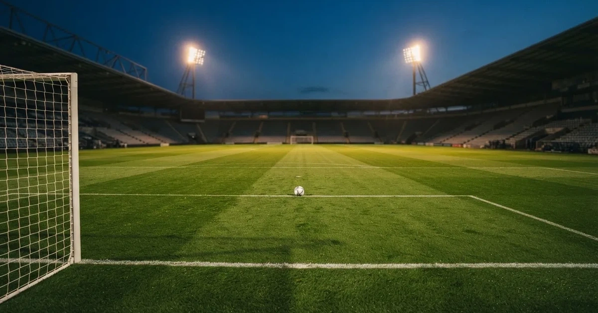 Ein Rasenplatz in der Dämmerung mit eingeschalteten Flutlichtmasten, ein Ball liegt am Anstoßpunkt, die Tribünen im Hintergrund sind unscharf.