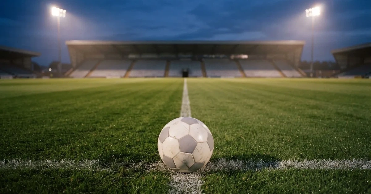Ein Fußball liegt auf dem Anstoßpunkt eines leeren Rasenplatzes, im Hintergrund eine Tribüne und Flutlichtmasten in der Abenddämmerung.