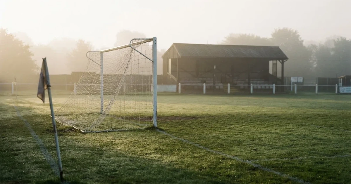 Ein Fußballtor mit Netz steht im Morgennebel auf einem leeren Rasenplatz, im Hintergrund die Silhouette einer kleinen Tribüne.