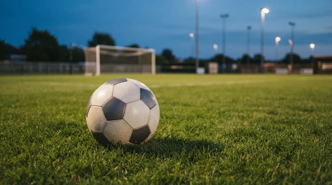 Ein Fußball liegt im Gras eines leeren Platzes, im Hintergrund stehen Flutlichtmasten in der Dämmerung.