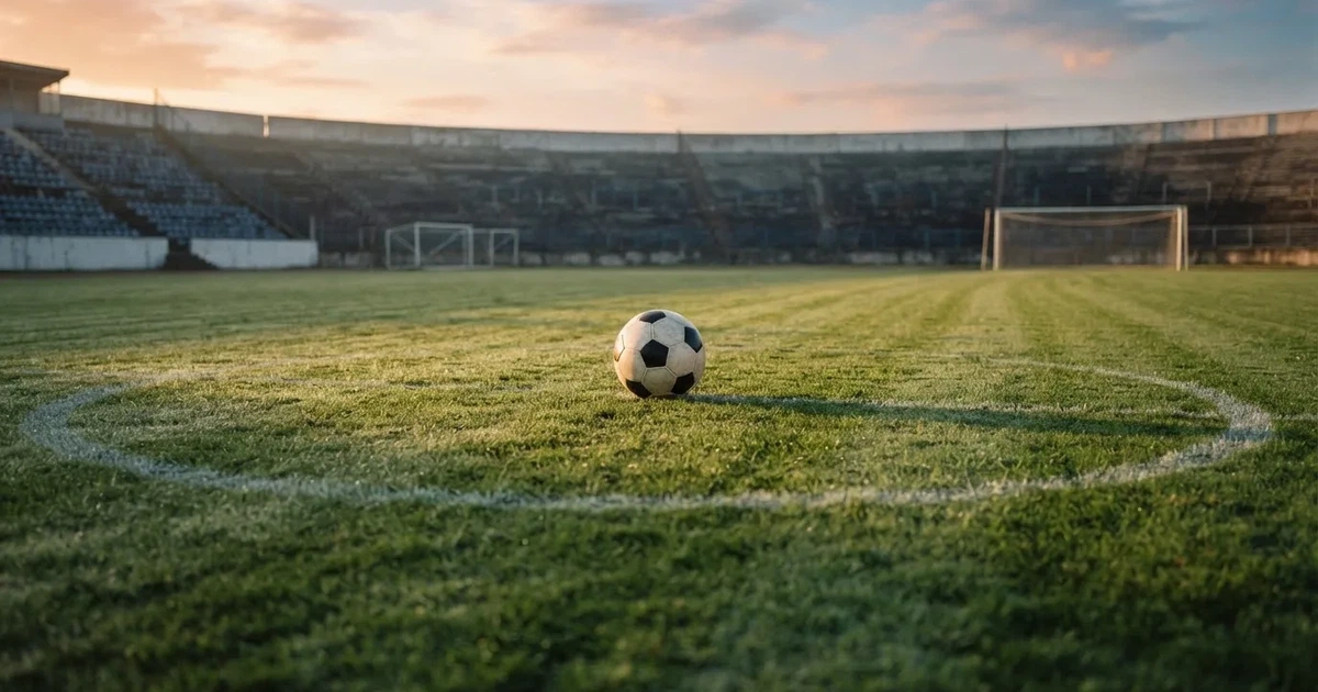 Ein Fußball liegt auf dem Anstoßpunkt eines Rasenplatzes, dahinter leere Tribünen im Licht des frühen Morgens.