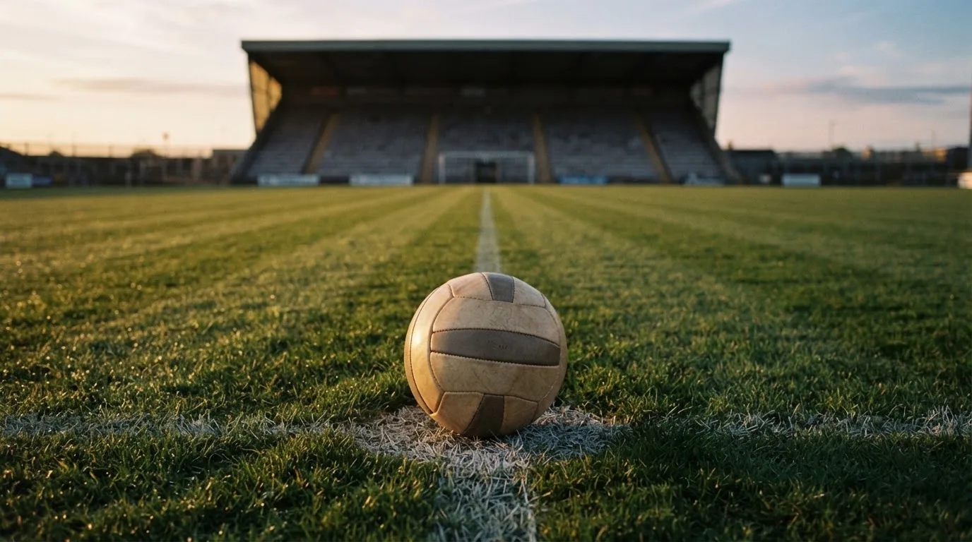 Ein Fußball liegt auf dem Anstoßpunkt eines Rasenplatzes, dahinter eine leere Tribüne im Abendlicht.