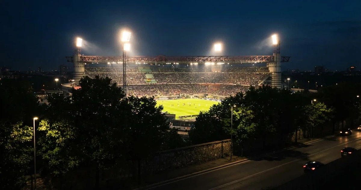 Ein Fußballstadion bei Nacht aus großer Entfernung, hell erleuchtet unter Flutlicht, davor eine dunkle Straße und Bäume.