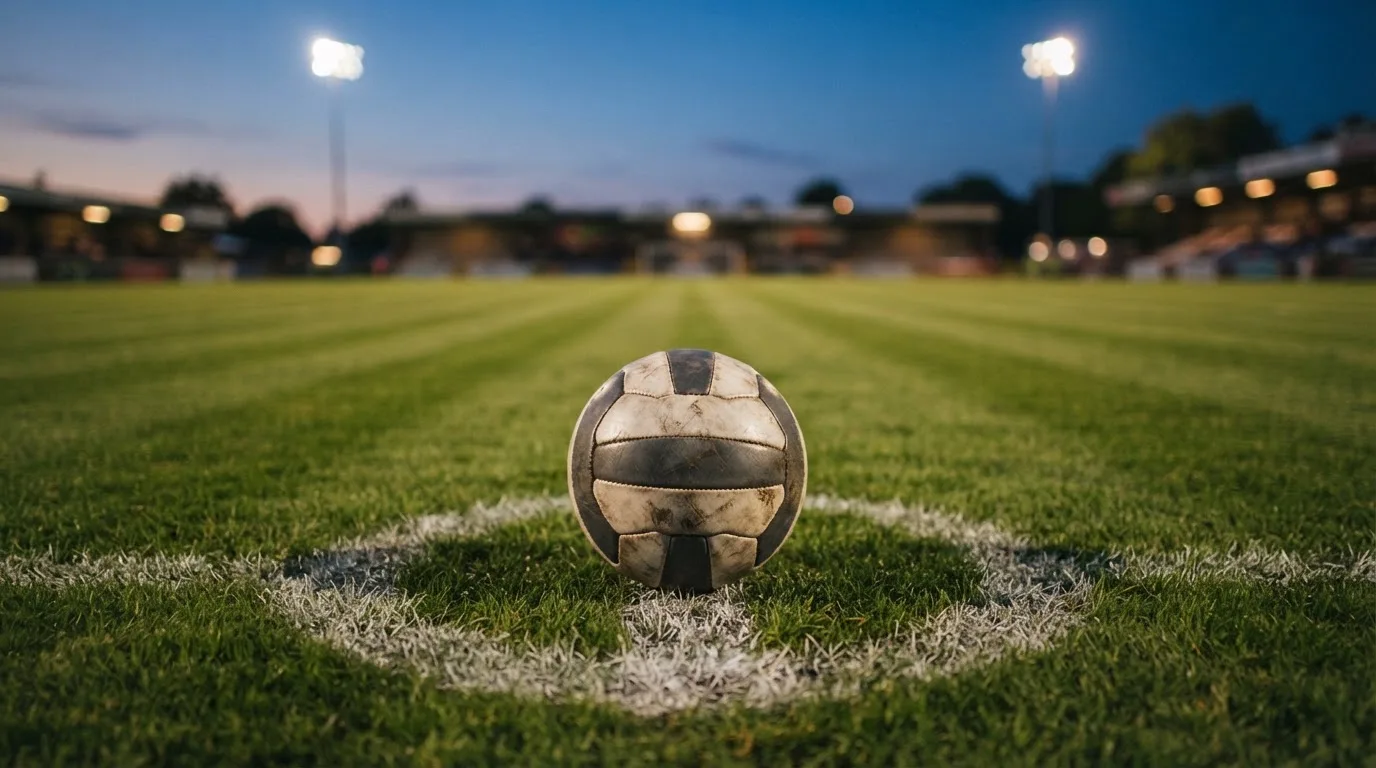Ein Fußball liegt auf dem Anstoßpunkt eines Rasenplatzes in der Abenddämmerung, Flutlichtmasten stehen unscharf im Hintergrund.
