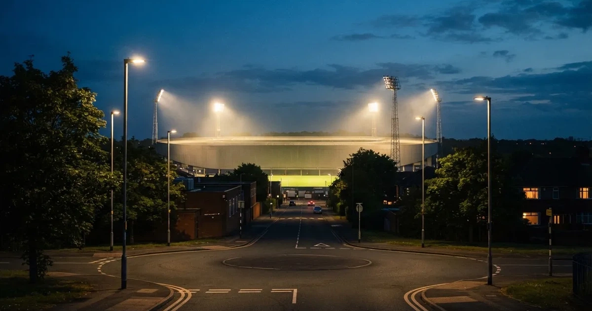 Ein Fußballstadion in der Dämmerung aus der Ferne, die Flutlichter gerade eingeschaltet, davor ein Kreisverkehr mit Laternen.