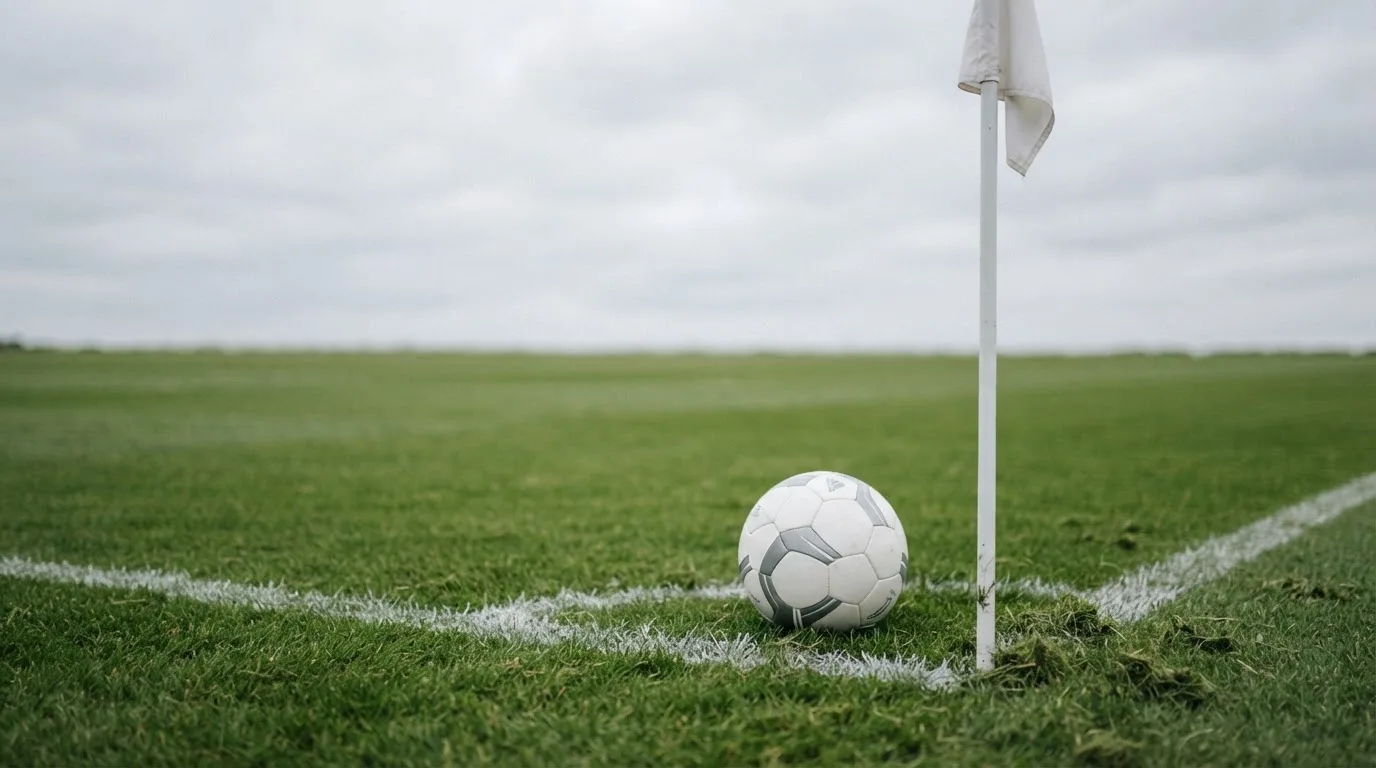 Ein Fußball liegt neben einer Eckfahne auf kurz gemähtem Rasen unter bewölktem Himmel.