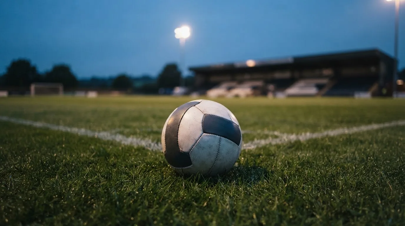 Ein Fußball liegt auf einem Rasenplatz in der Dämmerung, im Hintergrund ein ferner Stadionrand unter Flutlicht.